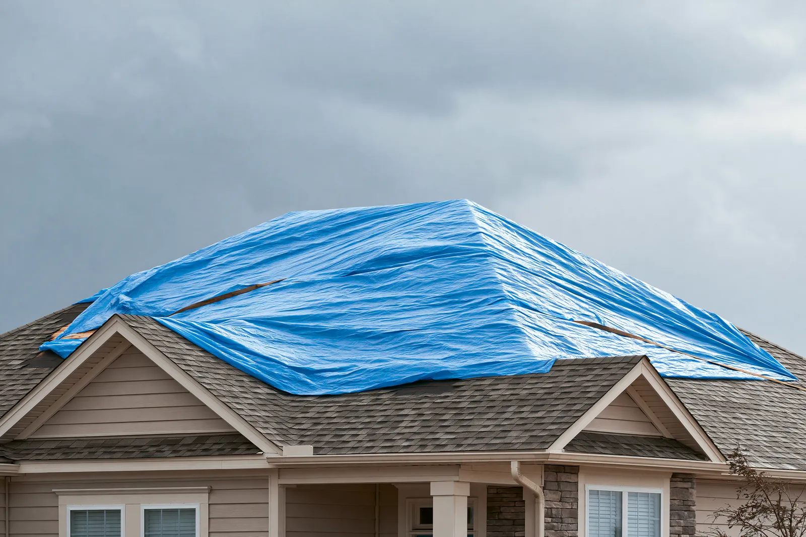 A two-story home's storm-damaged roof sections lie covered by a blue tarp under grey clouds